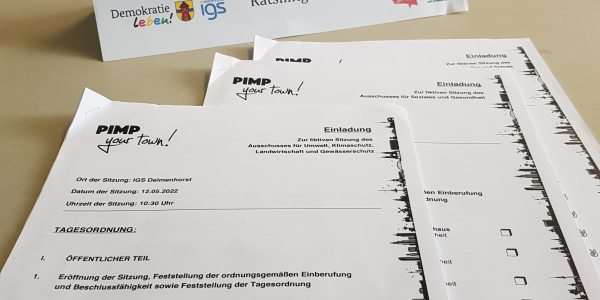 Auf dem Bild ist ein Namenssschild zu sehen, das die Symbole der Stadt Delmenhorst, der IGS Delmenhorst und des Vereins Politik zum Anfassen e.V. zeigt. Außerdem sind drei aneinandergeheftete Zettelblöcke zu erkennen, die Einladungen und Tagesordnungen zu